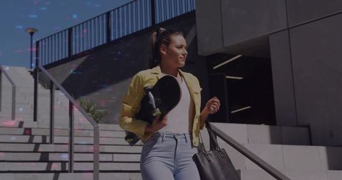 Young woman descending urban stairs carrying skateboard and tote bag in sunny city