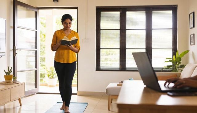 Woman reading while walking on yoga mat in sunlit modern living room, mustard top