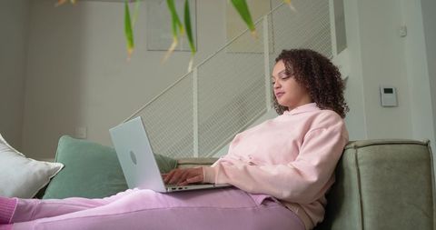 African american woman working remotely on laptop in pink loungewear on cozy sofa at home