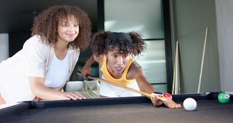 Female friends leaning and lining up precise pool shot on black-felt table