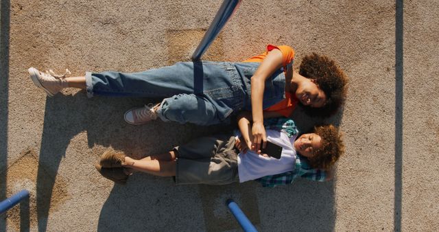 Two Generation Z siblings lying on outdoor ground in sunlight enjoying their time together. Perfect for themes highlighting sibling relationships, carefree youth, and outdoor leisure activities. Could also be used in campaigns emphasizing the use of technology and social media among young generations.