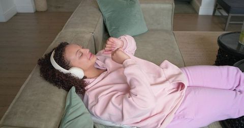 Reclining woman listening to music in pink hoodie on cozy sofa in living room