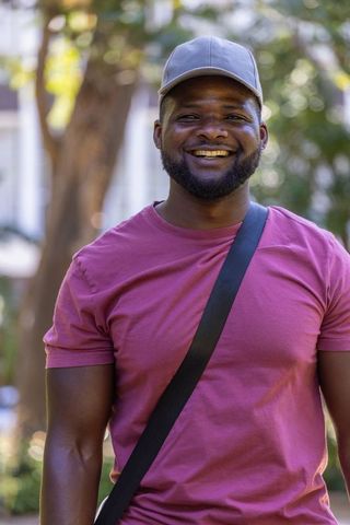 Smiling african american man standing in sunlit park wearing gray cap and crossbody bag