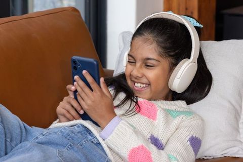 Indian girl laughing with headphones on sofa using smartphone cozy casual home moment