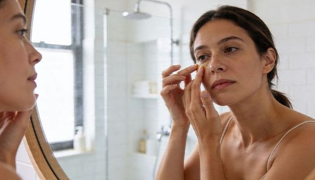 Woman applying under-eye cream in minimalist bathroom mirror morning skincare routine