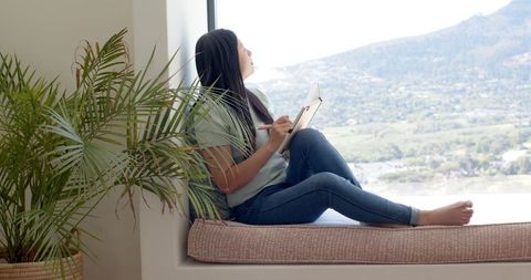 Young woman writing in journal on window seat overlooking valley and mountain view