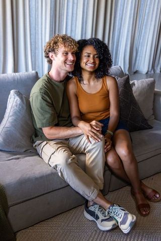 Young interracial couple sitting closely on light gray sofa holding hands and smiling warmly