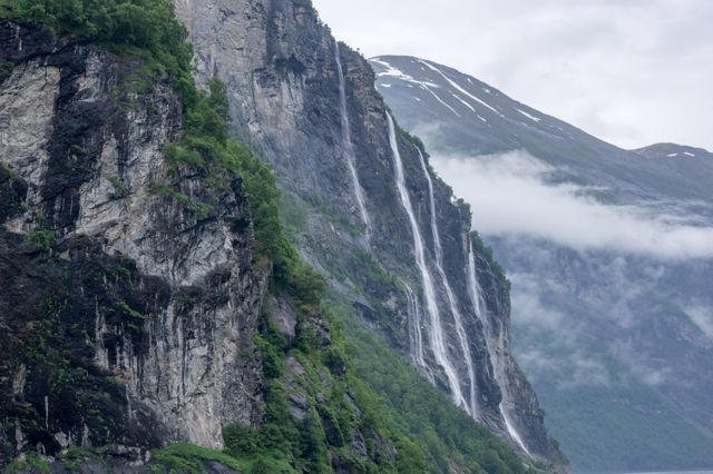 Majestic waterfalls cascade down rugged mountain cliffs, flowing into a serene fjord below. Mist envelops the scene, adding a mystical touch to the lush greenery of the forested slopes. This image can be used for travel promotions, nature documentaries, or environmental projects highlighting natural beauty and scenic landscapes.