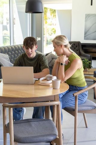 Mother and teenage son collaborating on laptop at home dining table in bright modern interior
