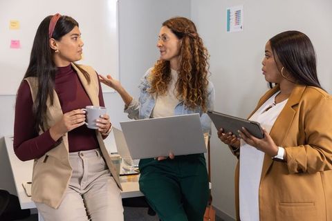 Diverse female team collaborating in modern office holding laptop, tablet and coffee mug