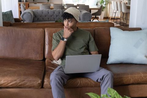African american young man working remotely on laptop while thinking on leather couch