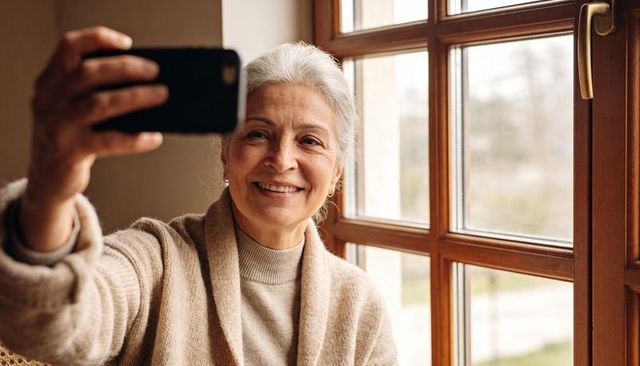 Senior woman smiling while taking selfie with smartphone by sunlit wooden window in cozy cardigan
