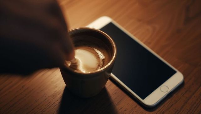 Stirring latte foam beside smartphone on wooden table, warm cozy coffee moment