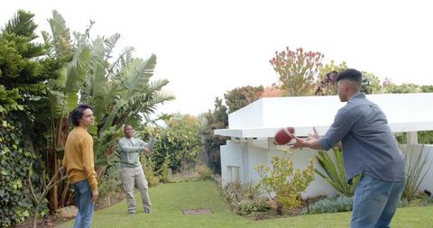Three diverse friends playing backyard catch with american football on grassy lawn