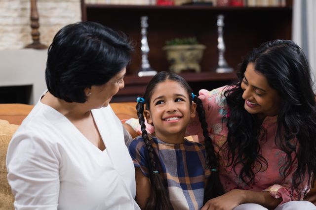 Three generations of women sitting together on a sofa at home, with a young girl smiling happily between her mother and grandmother. Perfect for use in family-oriented advertisements, parenting blogs, or articles about multigenerational living and family bonding.