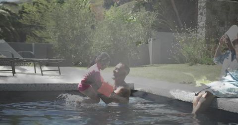 Smiling father lifting daughter in backyard pool, splashing during sunny summer day
