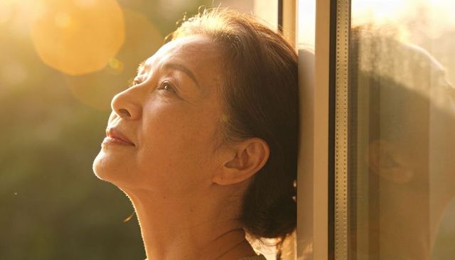 Sunlit asian mature woman leaning on window, reflective profile with golden lens flare