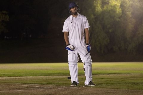 Late-night cricket batsman on floodlit pitch wearing helmet, pads and gloves, moody scene