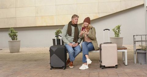 Mature couple waiting with rolling luggage on bench checking smartphone, planning trip