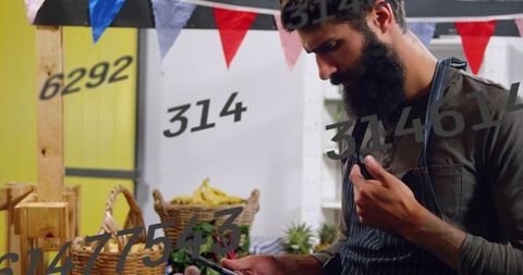 Bearded vendor checking smartphone while arranging bananas, greens at farmers market stall