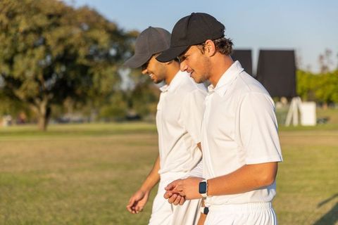 Sunlit cricket teammates walking across field in white kits and dark caps with scoreboard