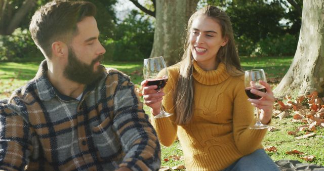 Couple sitting on blanket in park, holding glasses of red wine, enjoying sunny autumn afternoon. Ideal for promoting outdoor leisure activities, relationship bonding, wine industry, and autumn-themed events.