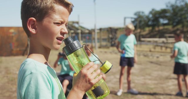 Young boy rehydrating during an outdoor physical activity on a warm sunny day. In the background, other children are participating in a fitness or sports activity, emphasizing themes of health, teamwork, and outdoor fun. Perfect for campaigns promoting kids' health and fitness, summer activities, or hydration awareness.