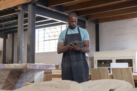 African american carpenter checking tablet and inspecting wooden planks in rustic workshop