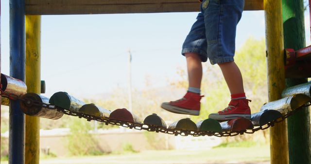 Young child wearing red shoes and jeans walking on colorful playground bridge. Ideal for themes of childhood joy, outdoor activities, and exploring. Great for use in educational materials, parenting blogs, and playground safety campaigns.