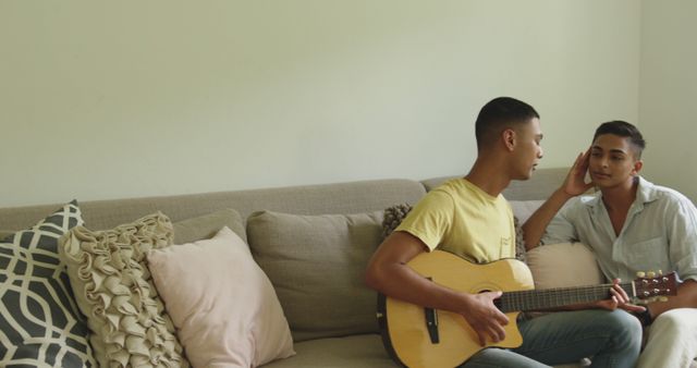 Biracial gay male couple sitting on sofa one man playing guitar the other listening. staying at home in isolation during quarantine lockdown.
