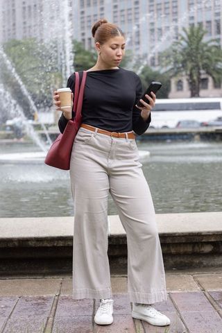 African american adult woman standing by fountain checking smartphone holding coffee red tote