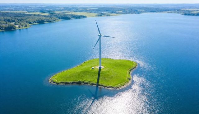 Wind turbine standing on green islet in blue lake casting long shadow for green energy