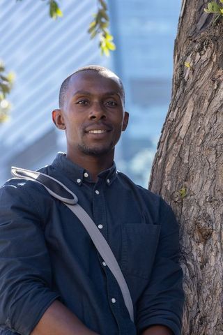 Confident african man leaning against tree near glass building, shoulder bag urban portrait