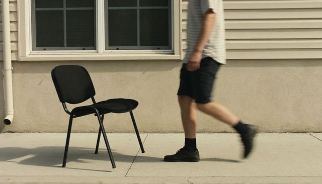 Man walking past empty black chair on sunlit sidewalk with building facade and strong shadows
