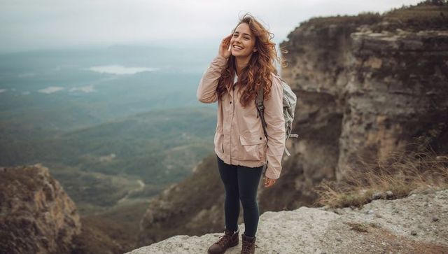 Smiling female hiker standing on cliff ledge overlooking expansive valley panorama overcast