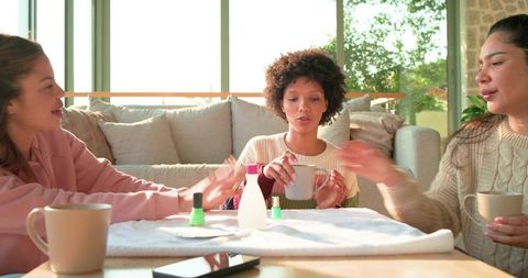 Female friends enjoying at-home manicure, sipping coffee, chatting in sunlit living room