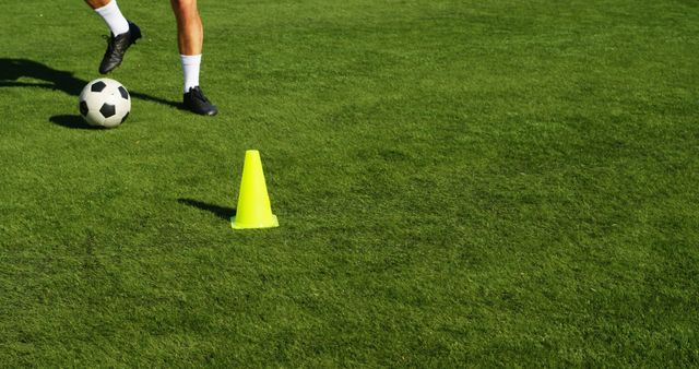 Soccer player dribbling ball near training cone on grass field. Useful for sports training articles, soccer skill tutorials, or promoting athletic wear and outdoor sports programs.