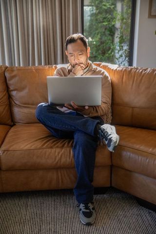 Asian man working on laptop while thinking on tan leather sofa in bright living room