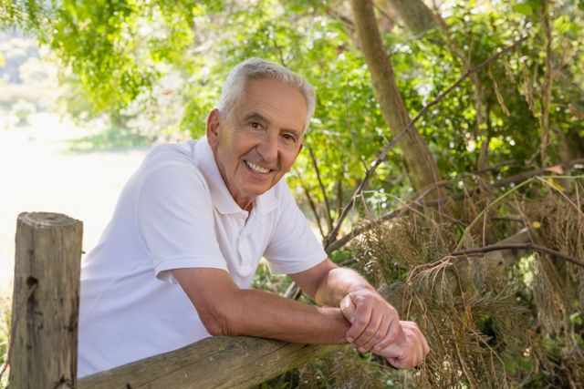 This image depicts a senior man leaning on a wooden fence in a forest, smiling warmly. Ideal for use in advertisements, articles, or websites related to retirement, senior living, outdoor activities, and healthy lifestyles for the elderly.