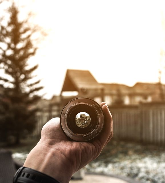 Useful for illustrating concepts of photography and technology, this stock photo features a hand holding a detachable camera lens in an outdoor setting. The lens captures the intricate details of the foreground while the background appears blurred. Ideal for articles, blog posts, and promotional material related to photography, visual techniques, or camera equipment.