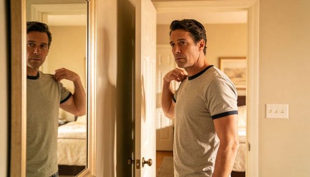 Midlife man adjusting t-shirt collar in bedroom doorway with mirror reflection
