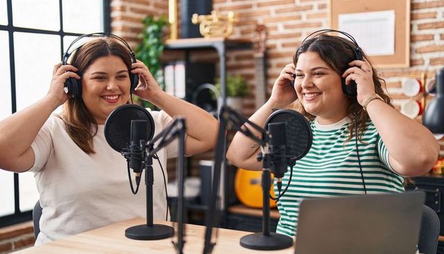 Latina cohosts recording podcast in cozy rustic studio adjusting headsets with laptop