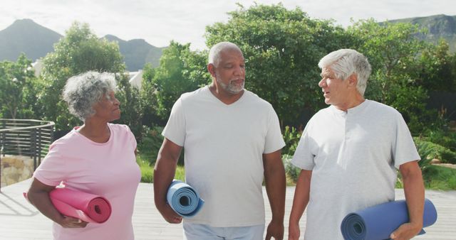 Three senior friends carrying yoga mats, walking outdoors on a clear day, looking excited and engaged in conversation. Perfect for use in promoting active lifestyles for older individuals, retirement community activities, or health and wellness programs focusing on fitness for seniors.