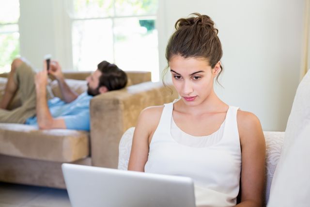 Beautiful young woman sitting on sofa using laptop with man in background