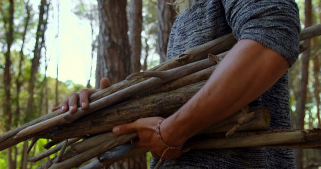 Person in forest gathering firewood by carrying several pieces of wood in arms. Image suitable for themes on outdoor activities, nature, survival skills, camping, and forest environments. Ideal for use in blogs, articles, and marketing materials related to nature exploration, outdoor living, environmental conservation, and adventure sports.