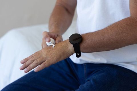 Mature man applying hand cream while wearing smartwatch on bed for everyday self-care