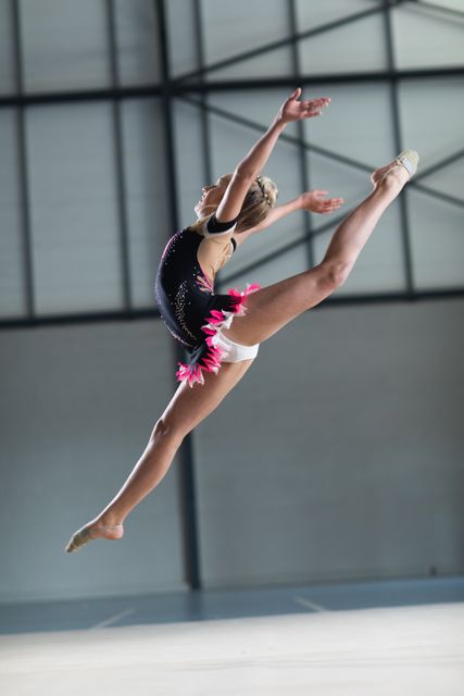 Caucasian female gymnast wearing a gymnastics costume practicing at the gym, jumping with arms and one leg raised in the air. Gymnast training hard for competition.
