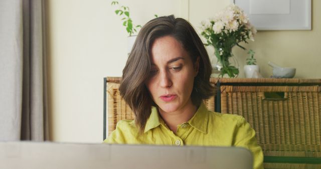 This image depicts a woman in a yellow shirt working intently on a laptop in a home office setting. Surrounding her are wicker baskets, a variety of potted plants, and light-toned walls, creating a serene atmosphere. This is ideal for use in articles or content related to remote work, productivity at home, freelance lifestyle, or creating a home workspace. It can also be used for themes related to professional women or work-life balance.