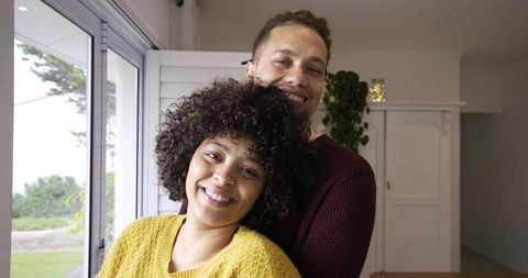 Smiling couple embracing at home near sliding glass door wearing cozy knit sweaters, relaxed
