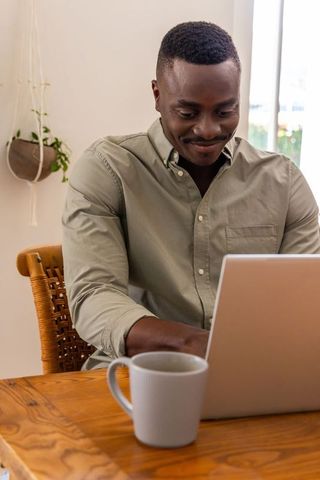 African american man working on laptop at home with coffee, natural light and greenery